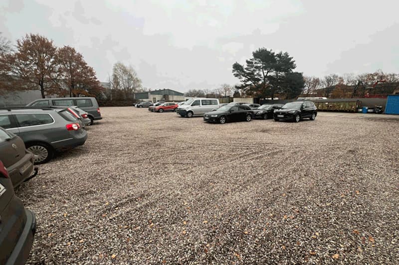 Großzügiger Schotterparkplatz mit verstreut stehenden Autos, Bäume und Lagergebäude im Hintergrund.