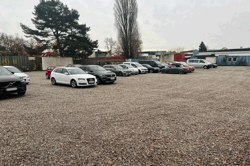 Schotterparkplatz mit mehreren geparkten Autos, Containerzaun und hohen Bäumen im Hintergrund.
