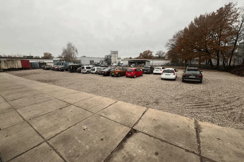Kiesparkplatz mit geparkten Autos, Betonplatten im Vordergrund und Industriegebäuden sowie Bäumen im Hintergrund.