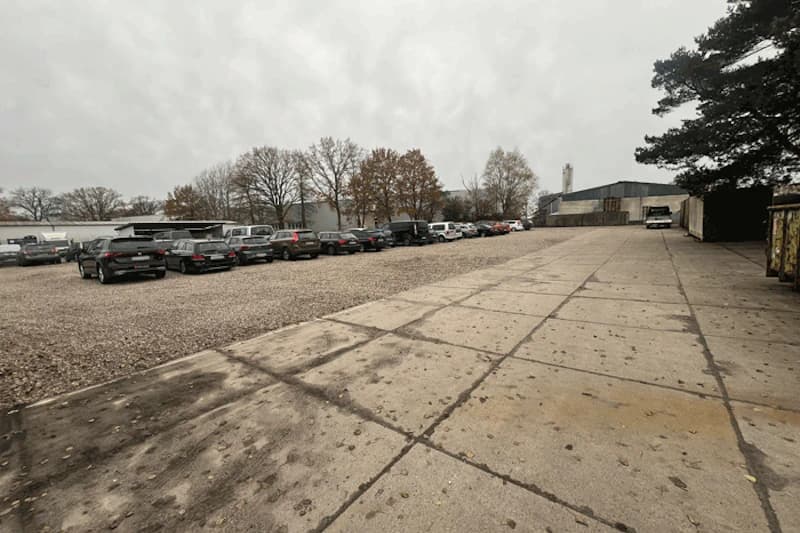Großer Außenparkplatz mit Reihen geparkter Autos auf Schotter und Betonplatten, Bäume und Hallen im Hintergrund.