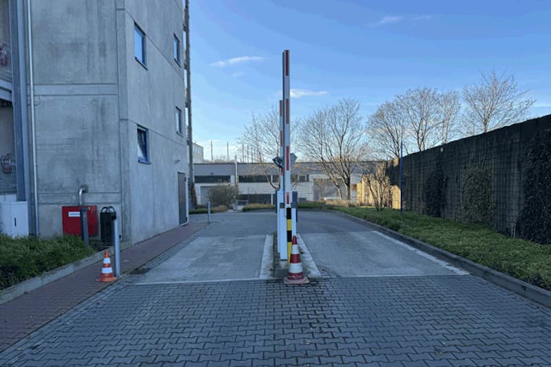 Schranke mittig zwischen zwei Fahrspuren, Verkehrsleitkegel, Betongebäude links und begrüntes Zaunelement rechts.