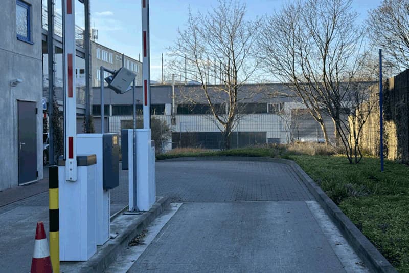 Zufahrt mit Schrankenanlage, zwei Kartenautomaten, Absperrkegel und Drahtzaun vor Hallenbauten.