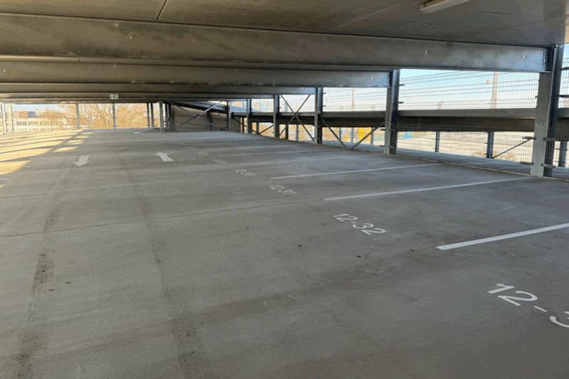 Oberes Parkdeck eines Parkhauses mit nummerierten Stellflächen, Betonboden, Stahlträgern und Gitterfencing.