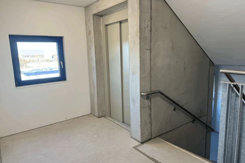 Treppenhausflur mit geschlossener Aufzugtür, Betonwänden, Handlauf und kleinem Fenster.