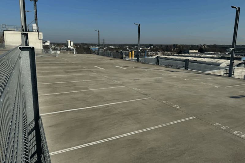 Leeres oberes Parkdeck mit markierten Parkflächen, Maschendrahtgeländer links und Laternen, Sicht auf Gebäude.