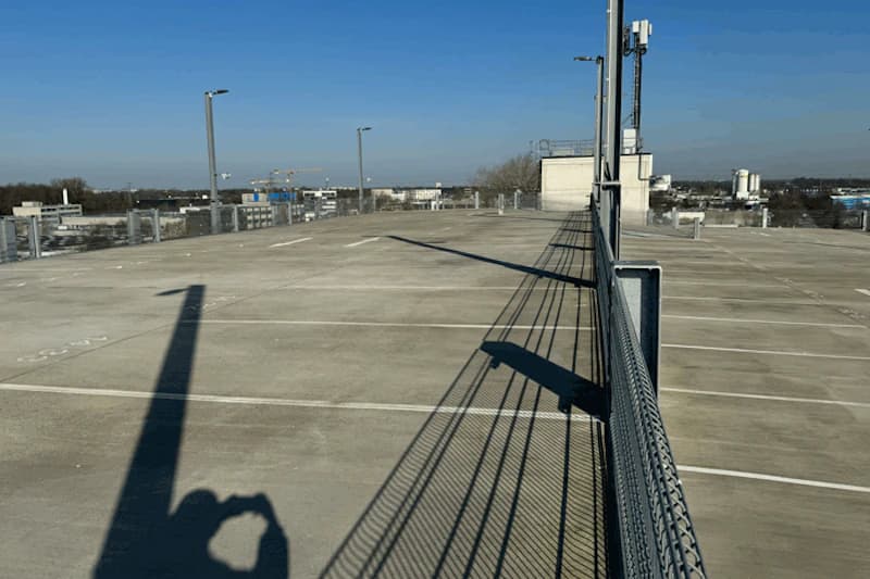 Leeres oberes Parkdeck mit Betonboden, Parkmarkierungen, Metallgitterzaun rechts und langen Schatten einer Person und Lampen.