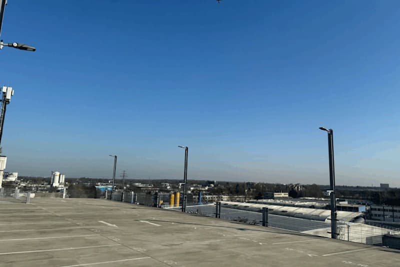 Leeres oberes Parkdeck mit Parkmarkierungen, Beleuchtungsmasten und Schutzgeländer, weiter blauer Himmel.