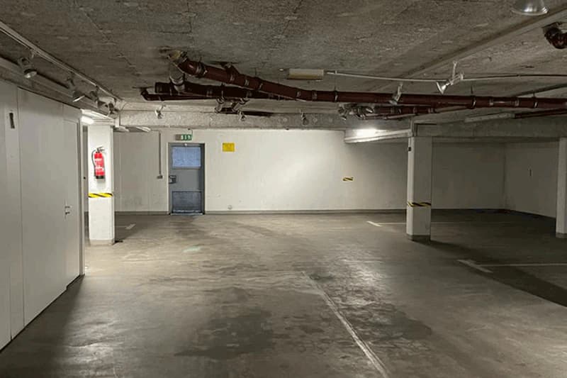 Leere Tiefgarage mit Stützpfeilern, sichtbaren Deckenrohren, Feuerlöscher und Notausgangstür.