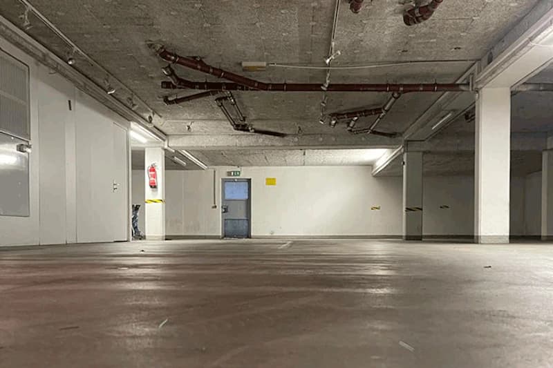 Leere Tiefgarage mit niedrigem Blickwinkel, glattem Betonboden, sichtbaren Deckenrohren und blauer Ausgangstür.