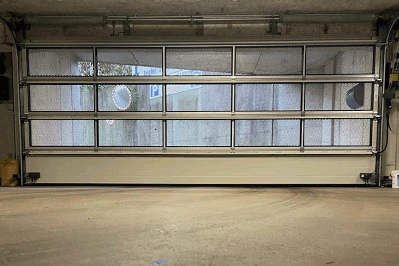 Geschlossenes Garagentor mit Metallgitter, dahinter Betonwand und Parkzeichen sichtbar.