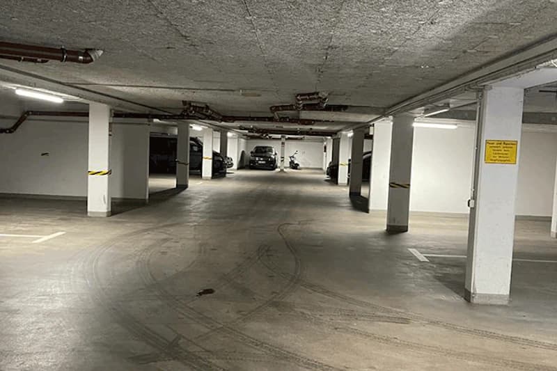 Unterirdische Tiefgarage mit Pfeilern, geparkten Autos im Hintergrund und Reifenspuren auf dem Betonboden.