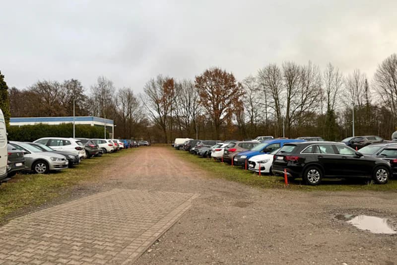 Außenparkplatz mit zwei Reihen geparkter Autos auf Schotterweg, Bäume im Hintergrund und grauer Himmel.