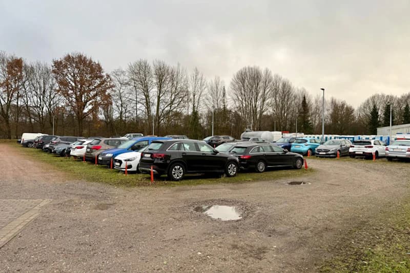 Mehrere Reihen geparkter Autos auf Schotterplatz mit Pfützen, orange Pfähle und kahle Bäume unter bewölktem Himmel.