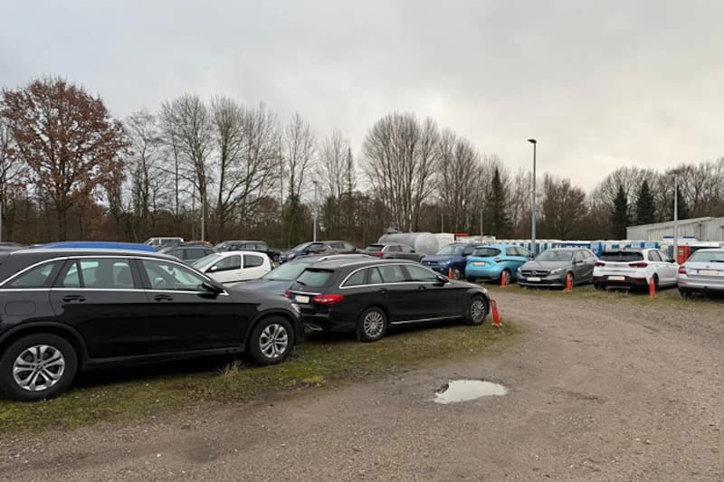 Freiluft-Parkplatz mit zahlreichen geparkten Autos auf unbefestigtem Untergrund und kahlen Bäumen im Hintergrund.