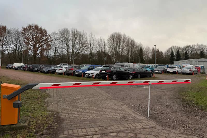 Geschlossene Schranke vor Freiluftparkplatz mit abgestellten Autos und kahlen Bäumen unter bedecktem Himmel.