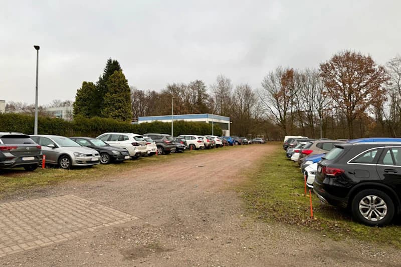 Offener Schotterparkplatz neben Bäumen und Hecke, Autos beidseitig geparkt, Laterne und kleines Gebäude im Hintergrund.