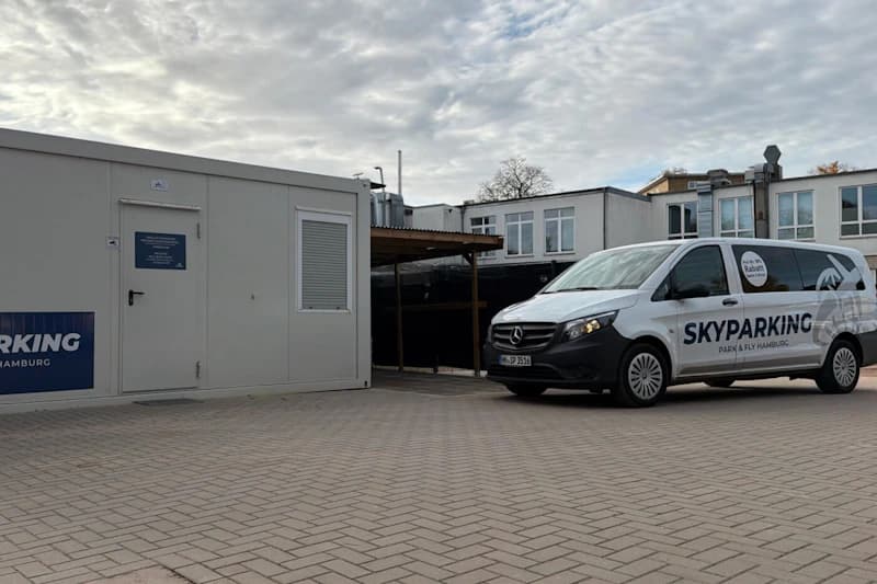 Gepflasterter Parkplatz mit weißem Containerbüro links und Skyparking Transferfahrzeug rechts unter bewölktem Himmel.