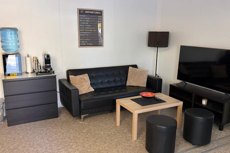 Aufenthaltsraum mit schwarzem Ledersofa, beigen Kissen, Holztisch, runden Hockern, großem Fernseher und Kommode mit Wasserspender.