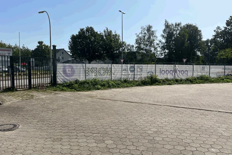 Gepflasterte Parkfläche mit Metallzaun, Werbebannern, Einfahrtstor und Laternen vor Baumbestand.