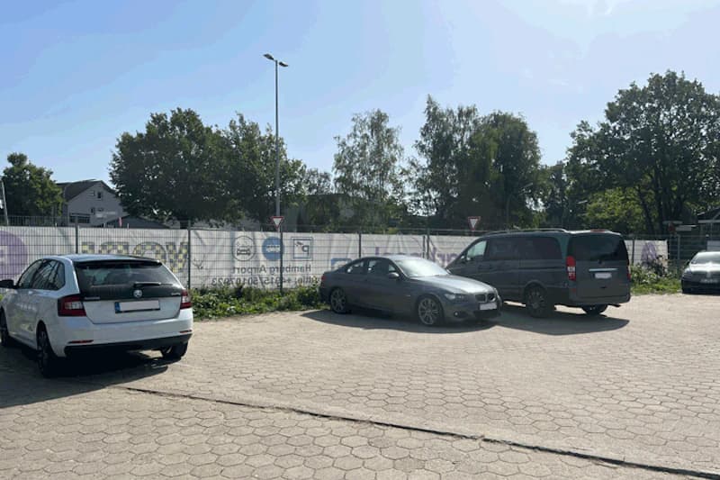 Gepflasterter, umzäunter Parkplatz mit drei geparkten Fahrzeugen, Laterne und Bäumen im Hintergrund.