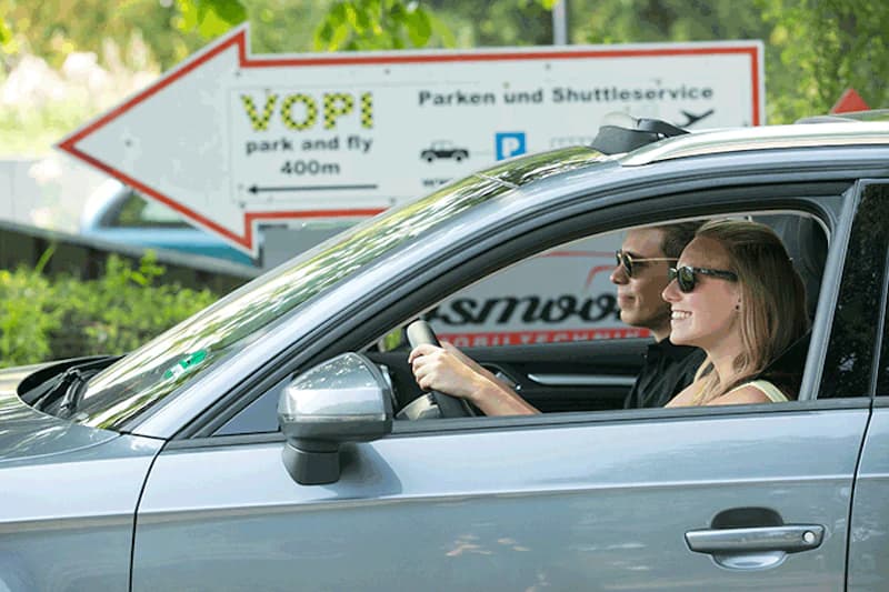 Zwei Personen sitzen lächelnd im Auto mit Sonnenbrillen, im Hintergrund ein Richtungspfeil mit VOPI-Schriftzug.