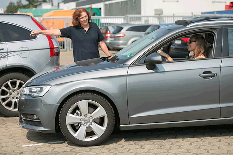Fahrerin im grauen Auto spricht mit einer stehenden Mitarbeiterin zwischen geparkten Fahrzeugen auf einem Parkplatz.