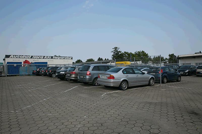 Gepflasterter Parkplatz mit mehreren geparkten Autos und umzäuntem Bereich neben Werkstatt.