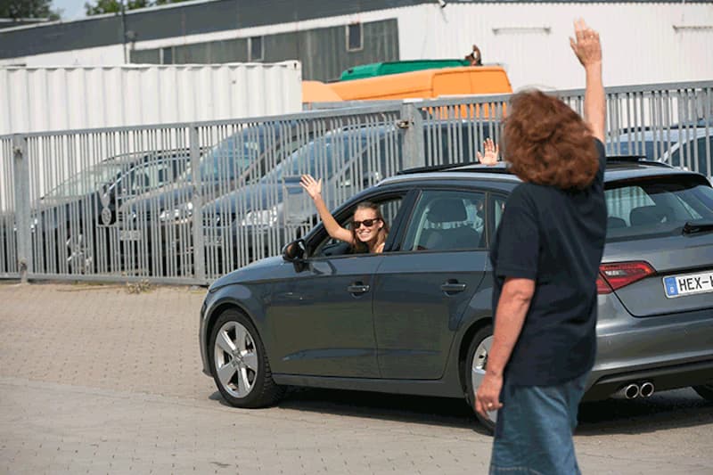 Graues Auto auf Parkplatz vor Metallzaun, Fahrerin winkt aus geöffnetem Fenster, eine stehende Person winkt zurück.