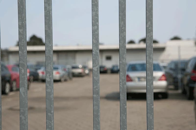 Sicht durch vertikale Metallstangen auf umzäunten Parkplatz mit geparkten Autos und Lagergebäude im Hintergrund.