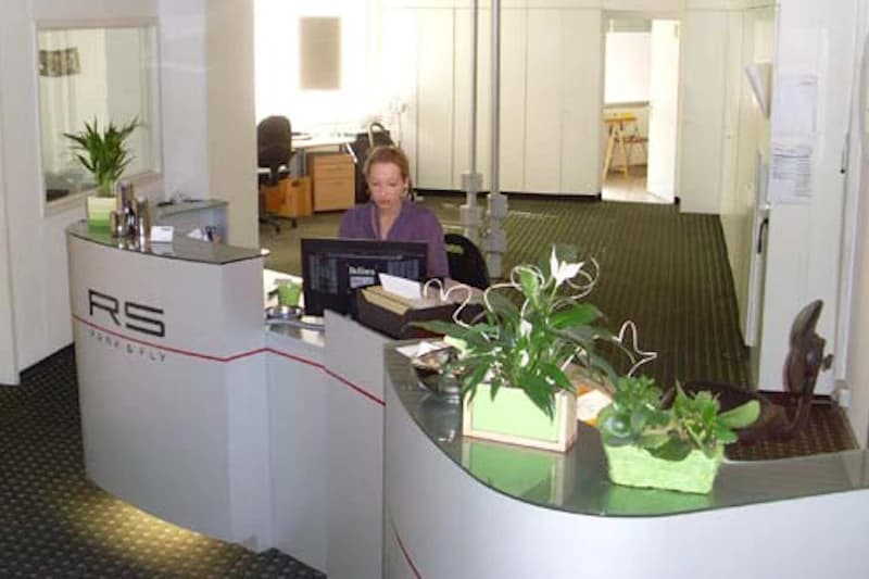 Empfangstheke mit Mitarbeiterin am Computer, Pflanzen auf der Theke und Büros im Hintergrund.