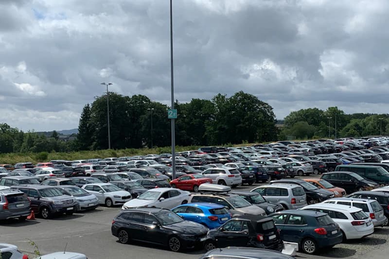 Großer Außenparkplatz mit vielen dicht geparkten Autos, Lampenmast mit Sektorzeichen und Wolkenhimmel.