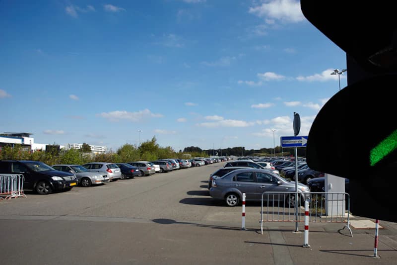 Großer Außenparkplatz mit Reihen geparkter Autos unter blauem Himmel, Verkehrsampel rechts mit grünem Licht.