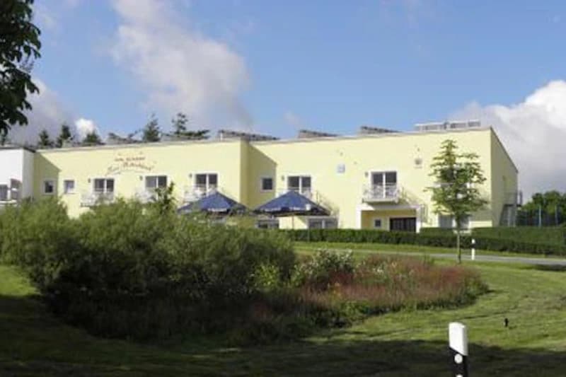 Gelbes zweistöckiges Hotelgebäude mit kleinen Balkonen, blauen Sonnenschirmen und Vorgarten mit Rasen und Büschen.