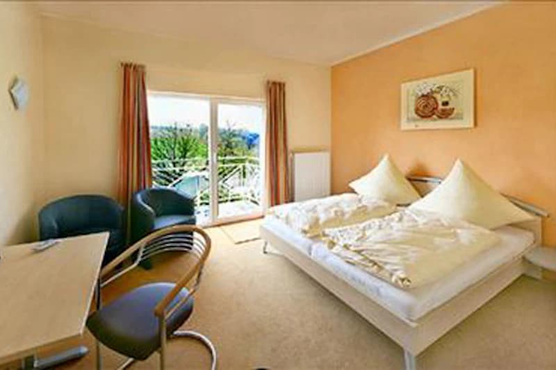 Schlafzimmer mit Doppelbett, zwei Sesseln, Schreibtisch und Balkon mit Glastür sowie orangefarbener Wanddekoration.