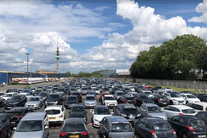 Großer Außenparkplatz mit dicht geparkten Autos in Reihen und Sendeturm im Hintergrund.