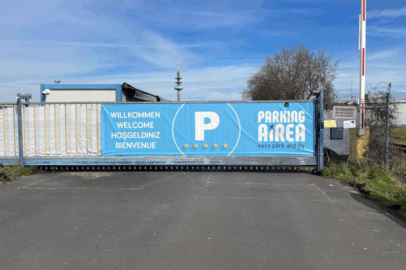 Verschlossene Schranke mit blauem Banner, Willkommensschrift und Parking Airea Logo am Flughafenzaun.