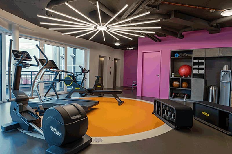 kleiner Fitnessraum mit Crosstrainern, Laufband, Hanteln, buntem Teppichkreis und großer sternförmiger Deckenleuchte.