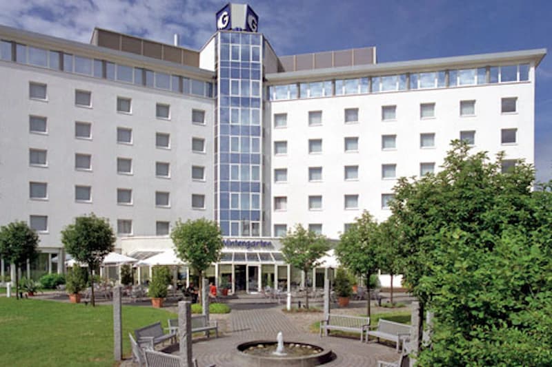 Weißes mehrgeschossiges Hotel mit mittigem verglastem Aufzugsturm, Vorgarten, Springbrunnen und Bänken.