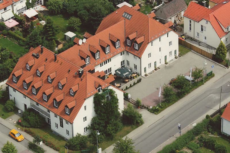Luftaufnahme eines weißen Gebäudes mit rotem Ziegeldach, vielen Dachgauben, Innenhof mit Sonnenschirmen und Parkplatz an der Straße.