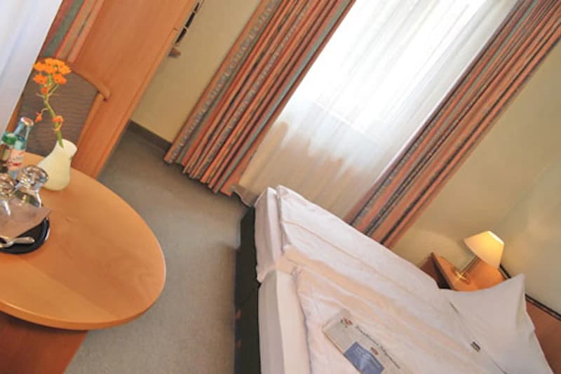 Helles Hotelzimmer mit zwei zusammengeschobenen Betten, Nachttischlampe, rundem Tisch mit Vase und Flasche sowie Fenster mit Vorhängen.