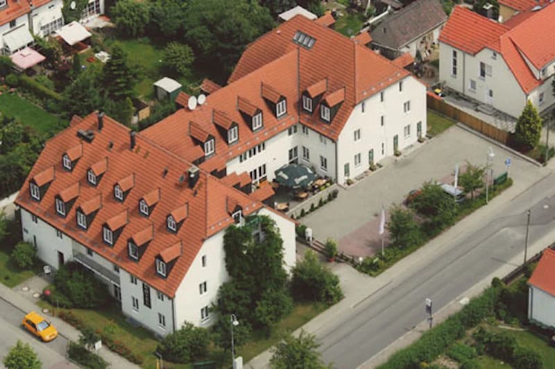 Weißes Hotel mit roten Ziegeldächern und vielen Gauben, Innenhof mit Tischen sowie Parkplatz an einer Straße.