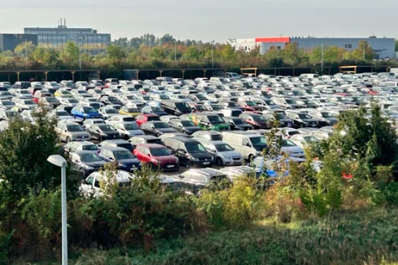 Großer Außenparkplatz mit dicht geparkten Autos, Grünstreifen im Vordergrund und Industriegebäuden im Hintergrund.