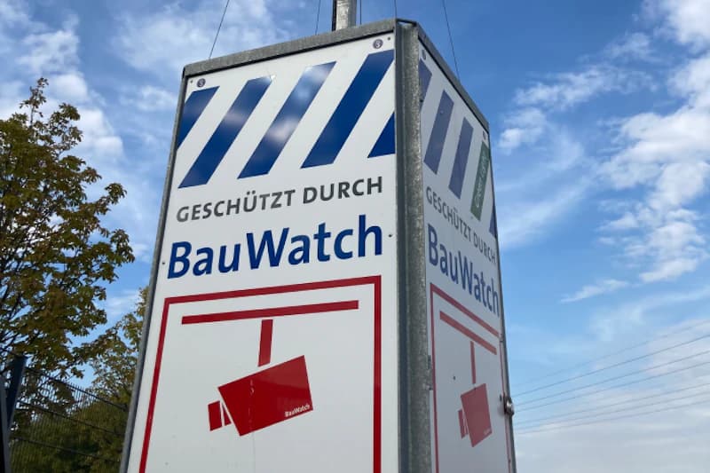 Metallschild mit Aufschrift Geschützt durch BauWatch und rotem Kamera-Piktogramm an einem Pfosten vor Himmel.