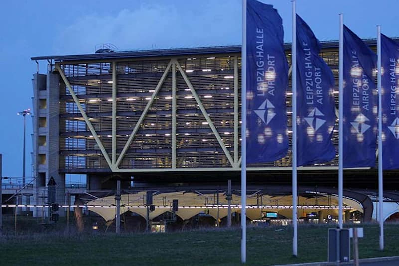 Mehrstöckiges Parkhaus mit diagonalem Stahlträgergerüst, beleuchteten Parkebenen und Fahnen mit Leipzig/Halle Airport im Vordergrund.