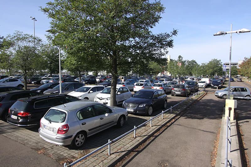 Außenparkplatz mit mehreren Reihen geparkter Pkw, Bäumen, Beleuchtungsmasten und Gehweg.