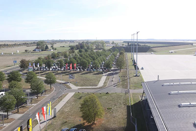 Übersicht über Parkplatz mit Baumreihen und geparkten Autos neben Flugfeld und Terminaldach.