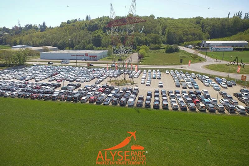 Großer Außenparkplatz mit Reihen geparkter Autos, Hochspannungsmast und umliegenden Feldern.