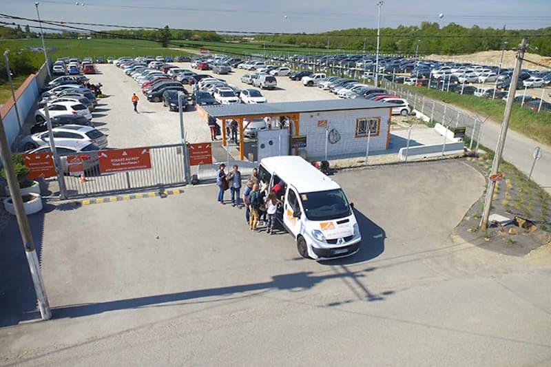 Abgegrenzter Parkplatz mit zahlreichen geparkten Autos, Schranke und Bürocontainer, Transferbus mit einsteigenden Personen am Eingang.