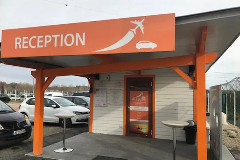 Kleiner orangefarbener Empfangspavillon mit Schild, zwei Stehtischen und mehreren geparkten Autos im Freien.
