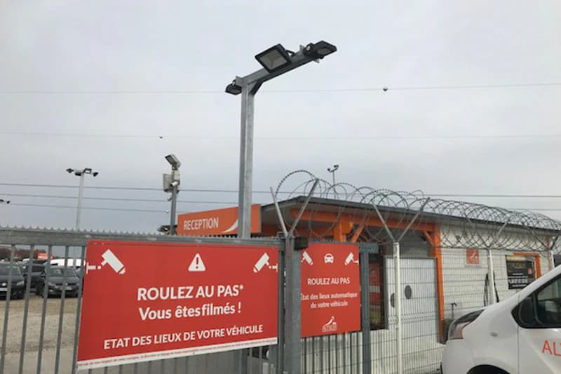 Gittertor mit Stacheldraht, roten Warnschildern und Überwachungskameras vor orangefarbener Empfangshütte.